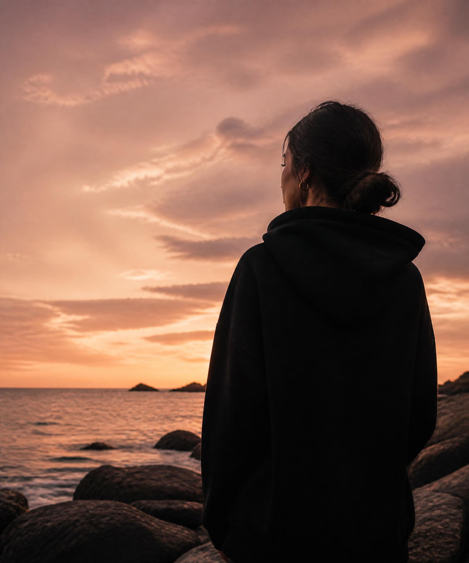 Woman in oversized black hoodie standing on a rocky shore at sunset